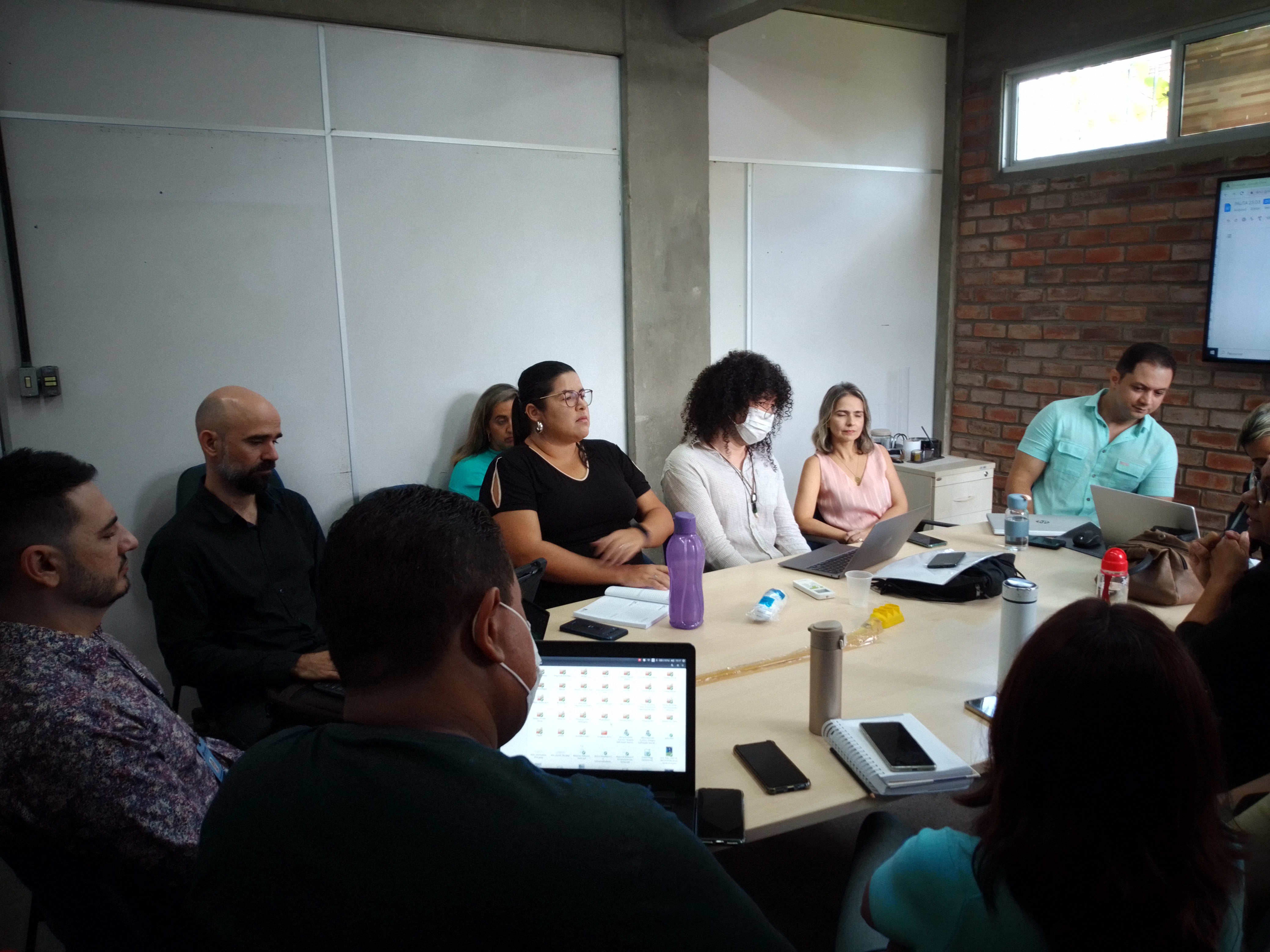 Pessoas sentadas ao redor de uma mesa, em uma sala. Sobre a mesa, estão laptops, cadernos, agendas, garrafas e copos.