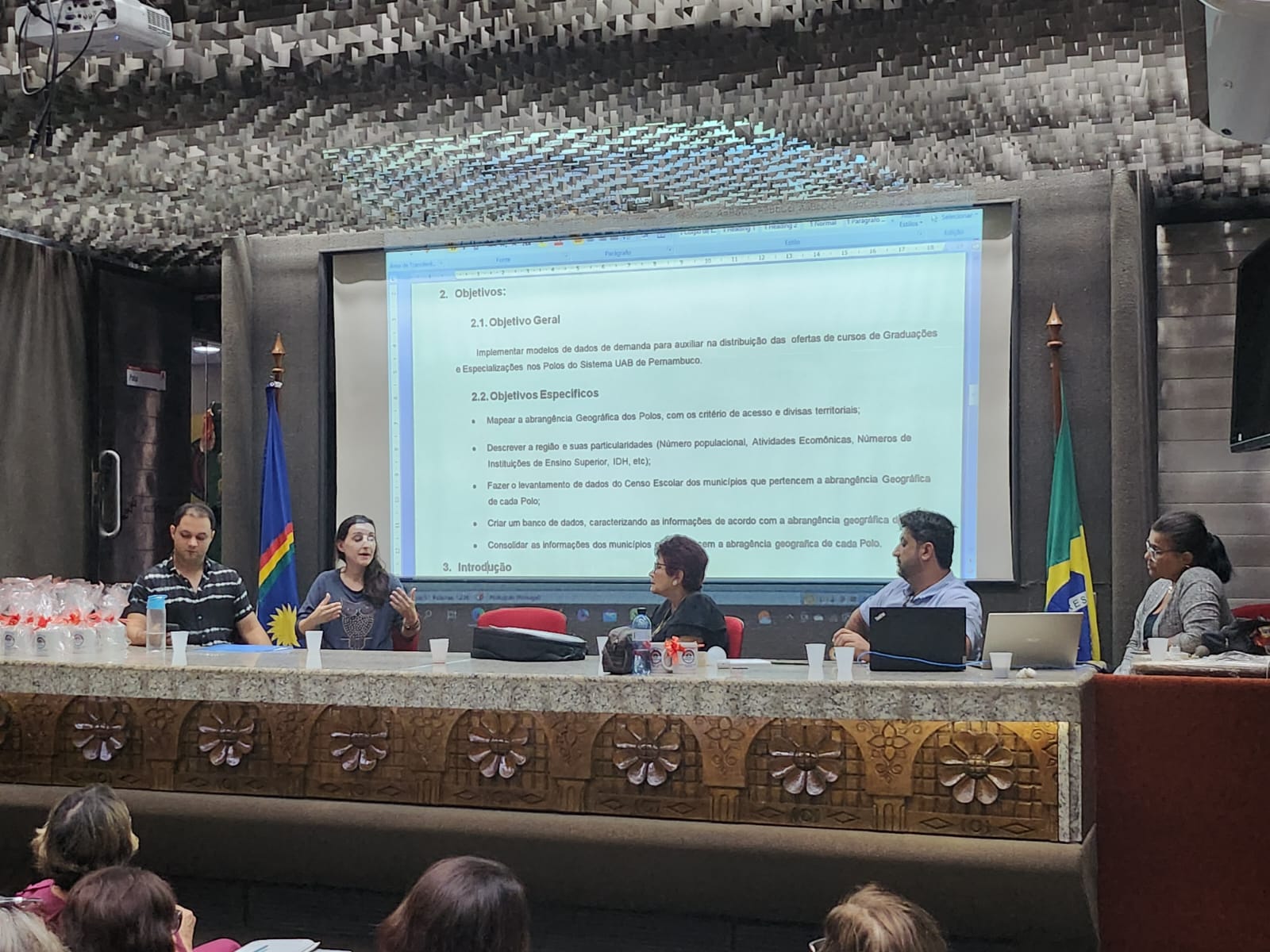 Foto da reunião de colegiado mostrando o Prof. Teo e a Prof. Elidiane sentados à uma mesa oval, em frente à uma grande tela de projeção, de frente para pessoas sentadas em uma plateia.  
